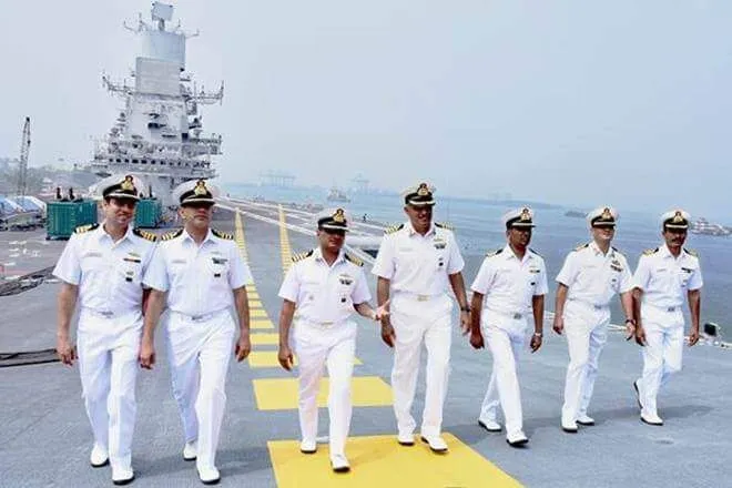 Seven naval officers in white uniforms and peaked caps walking together across the flight deck of an aircraft carrier