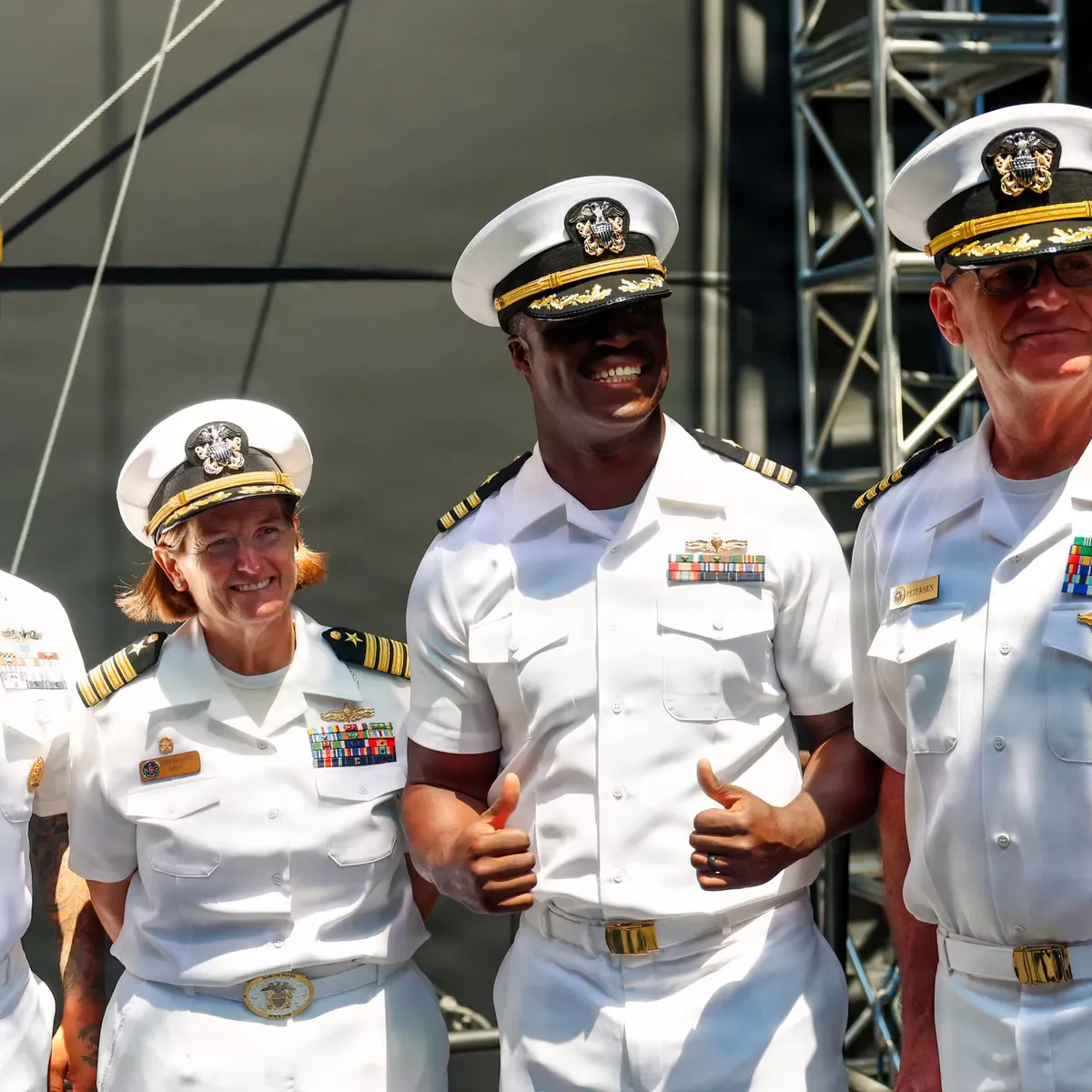 Three naval officers of different ranks in white dress uniforms smiling together at a public ceremony