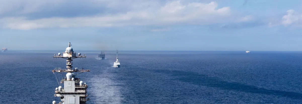 A ship’s island and radar mast above the open ocean with escort vessels on the horizon