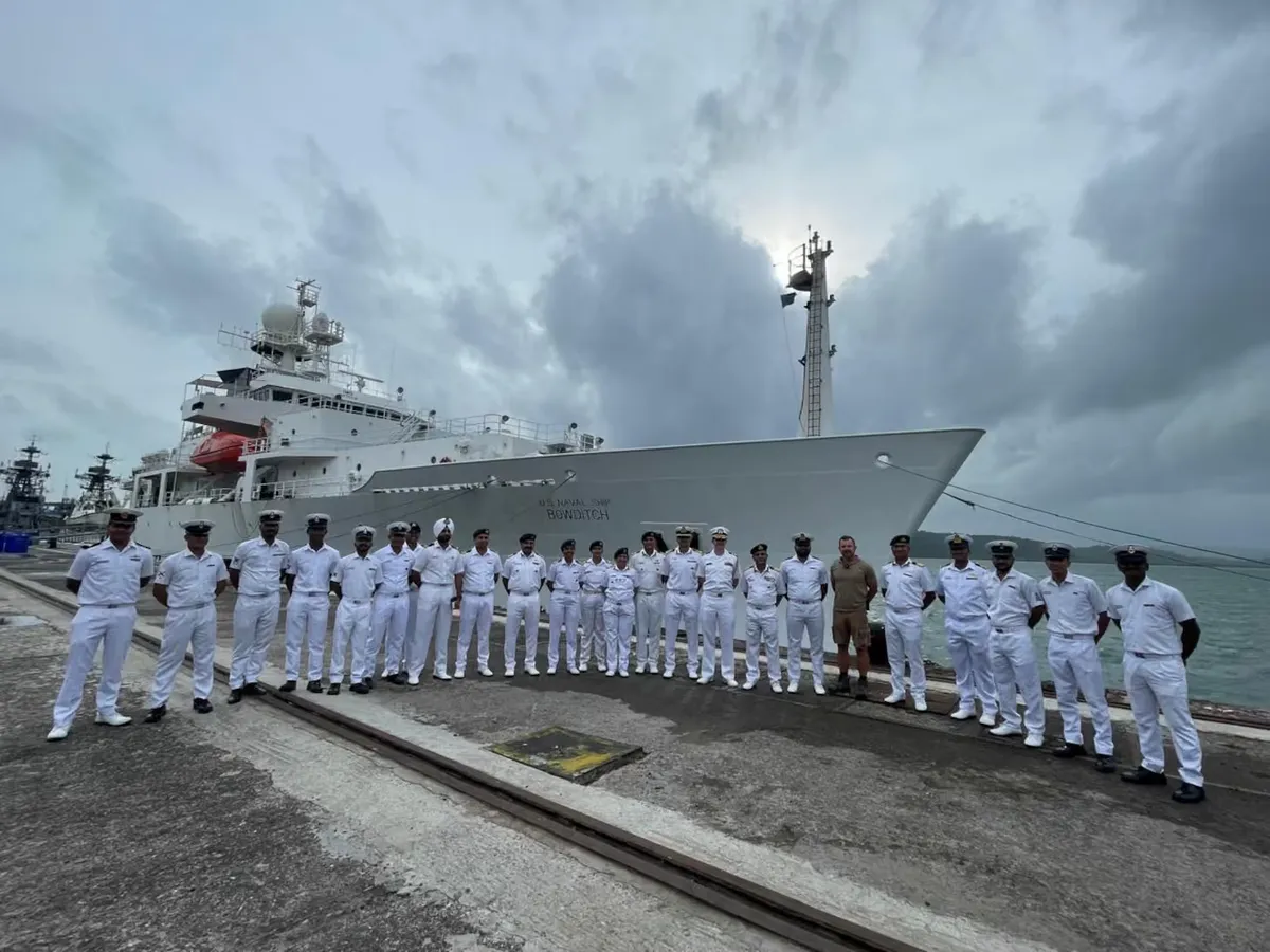 Naval personnel in white uniforms stand in a line on a quayside in front of a large survey ship, representing international naval workforce cooperation