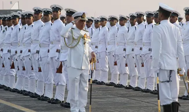 A senior naval officer salutes while reviewing long ranks of sailors in white uniforms at a ceremonial parade