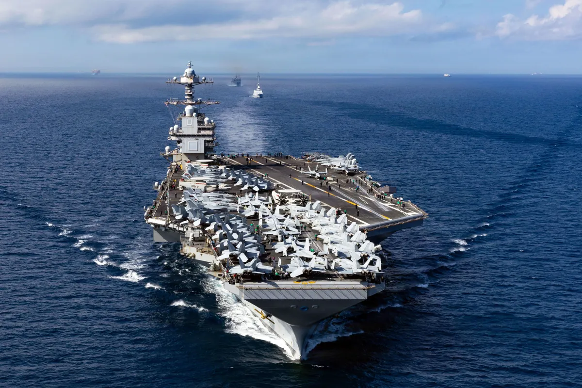 An aircraft carrier and escort vessels underway in formation across the open ocean, seen from above