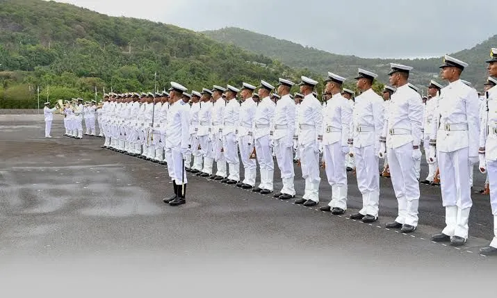 Ranks of naval trainees in white uniforms standing on a parade ground at a naval academy surrounded by green hills